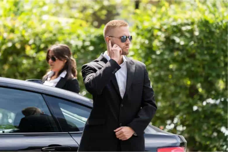 A man in a suit is standing next to a car, while engaged in a phone conversation about Newark Airport Transportation.