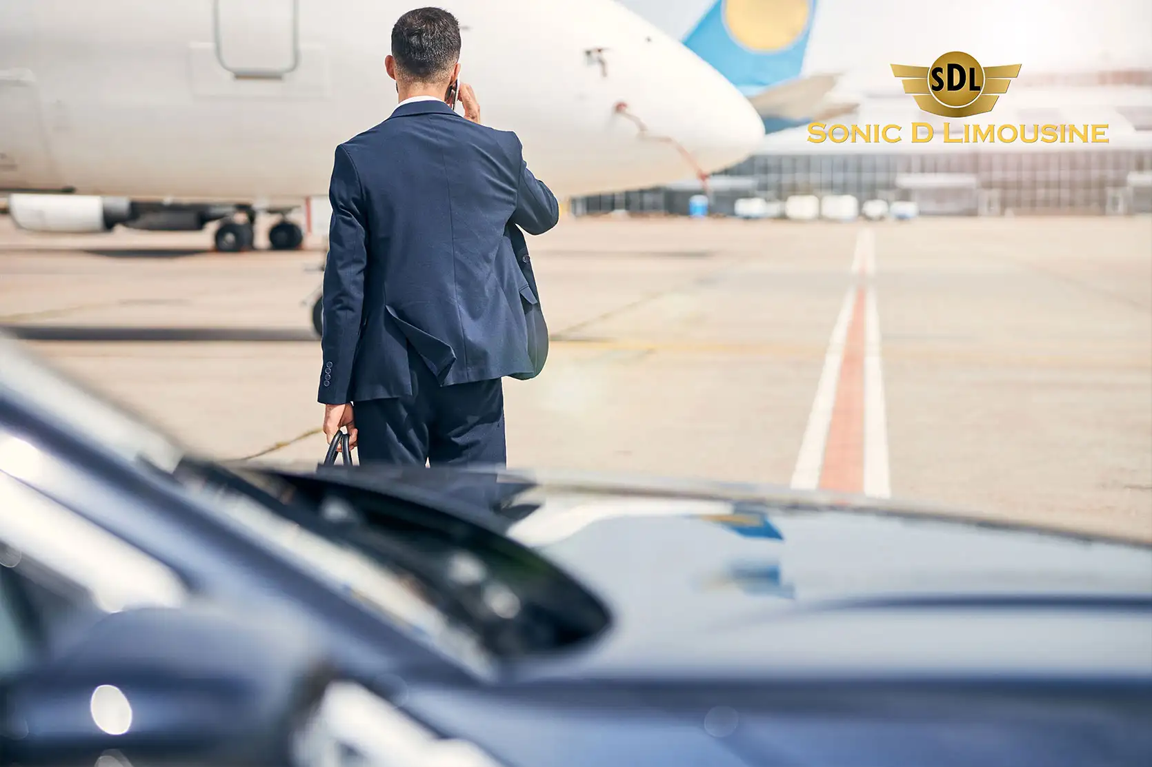 A man in a suit standing next to a car with an airplane in the background.